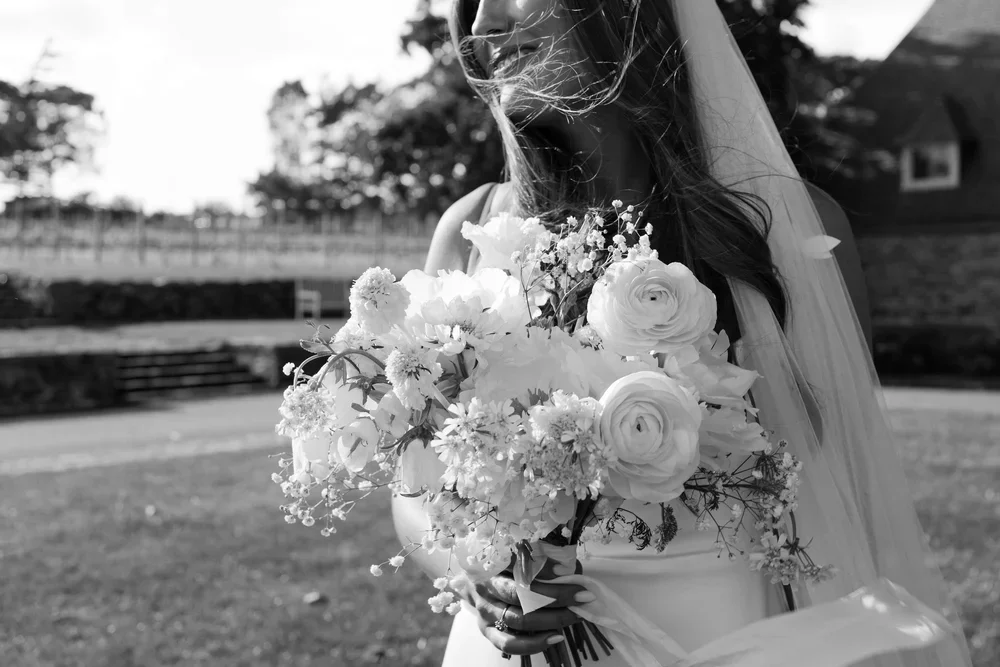 Black and white photo of a bride holding a large bouquet of flowers outdoors, with a veil and flowing hair, and a blurred background of trees and building.