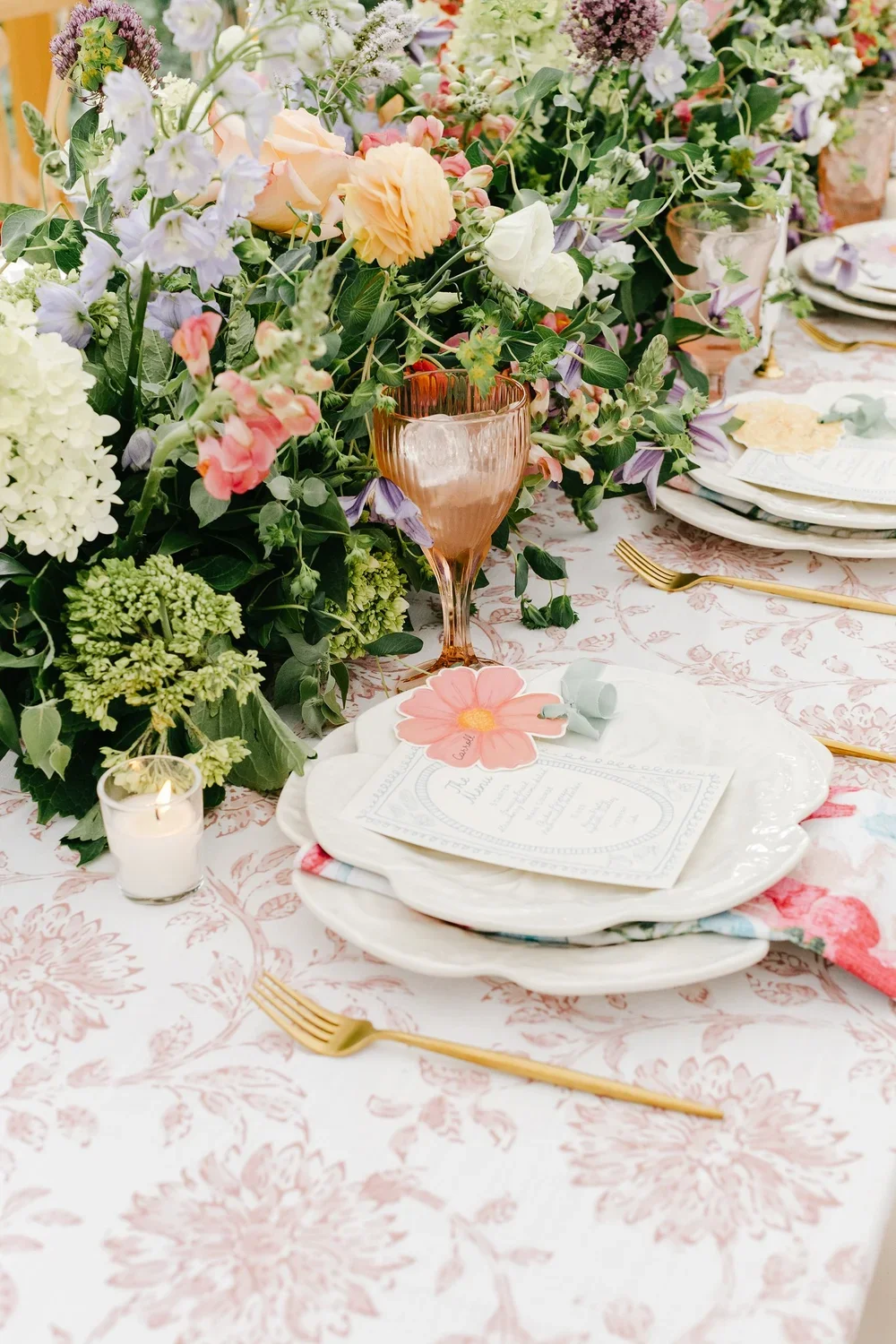 Elegant floral table setting with pink, purple, and white flowers, gold cutlery, pink glassware, a lit white candle, and layered white plates with a floral napkin and menu card.