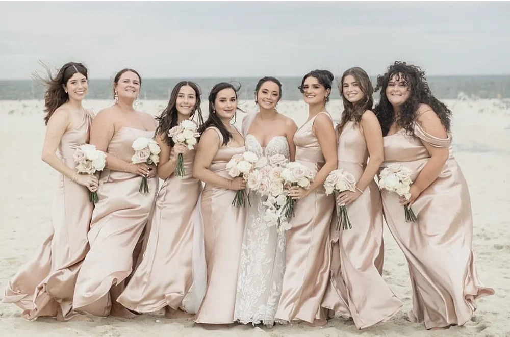 Bridal party standing on the beach, wearing matching blush pink dresses and holding bouquets of white flowers.