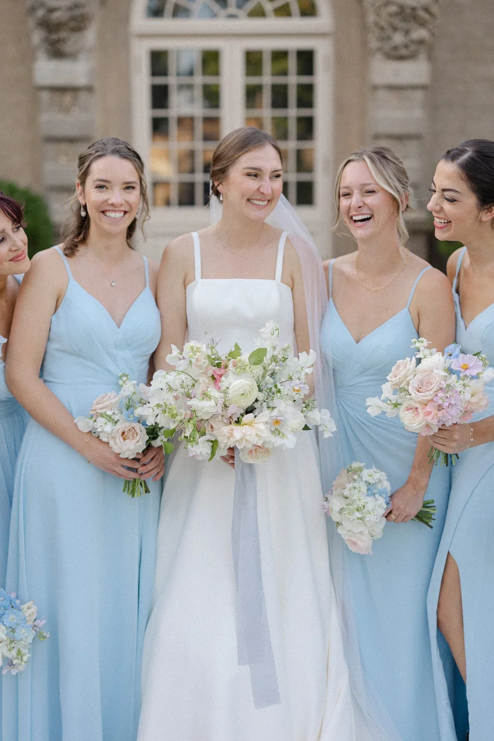 Bridal party with women in light blue dresses and a bride in white, holding bouquets, smiling outdoors in front of a building.