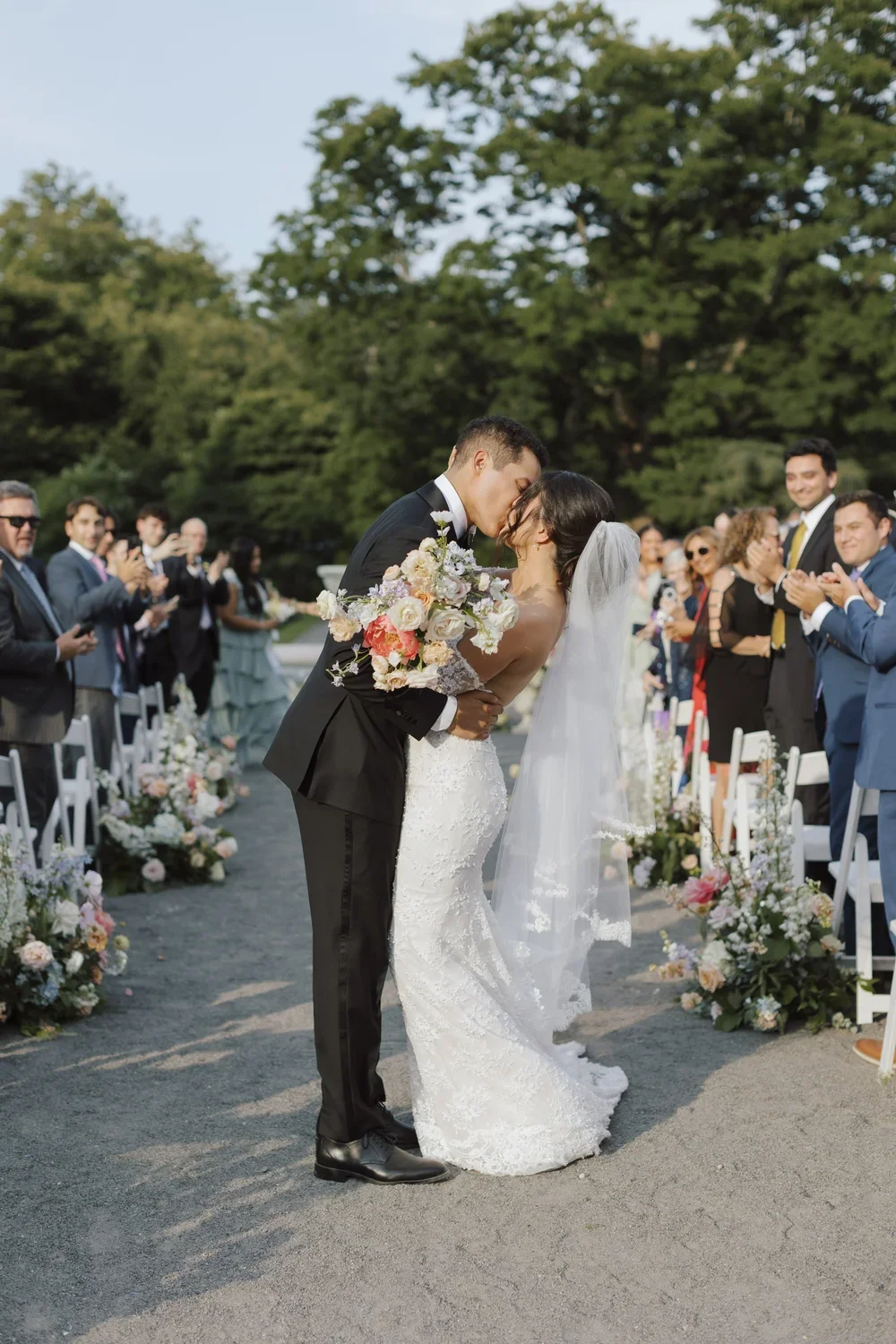 A wedding ceremony outdoors with a bride and groom kissing, surrounded by friends and family, with trees in the background.