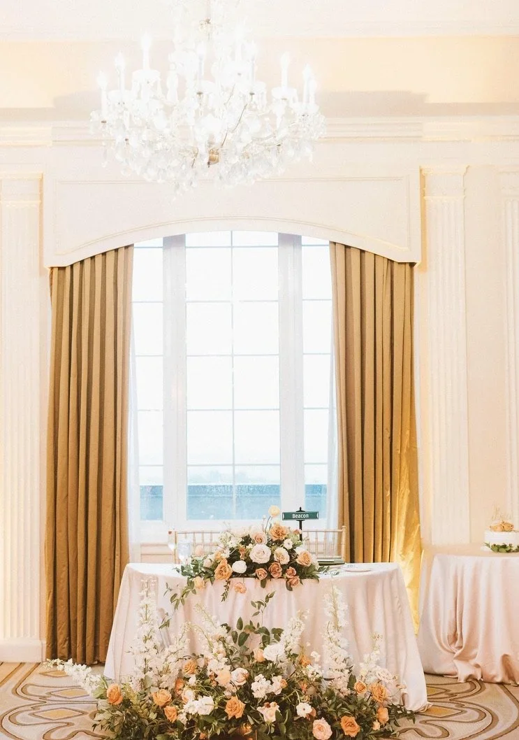 Elegant wedding or event reception table decorated with pink and white roses, greenery, and a floral arrangement, set in a room with large window, gold curtains, and a chandelier.