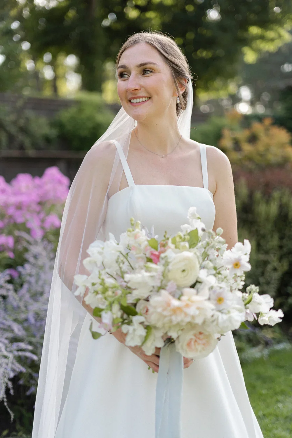 A smiling bride holding a bouquet of white and pink flowers in a garden setting.