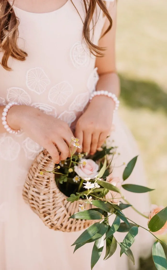 A young girl wearing a light-colored dress with white lemon slice embroidery, holding a small wicker basket filled with pink roses and greenery, outdoors on a bright day.