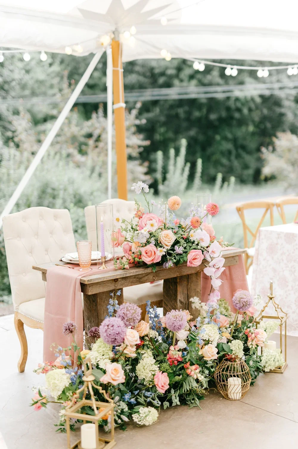 A decorated outdoor table with pink flowers, candles, and elegant tableware, set beneath a canopy with string lights.