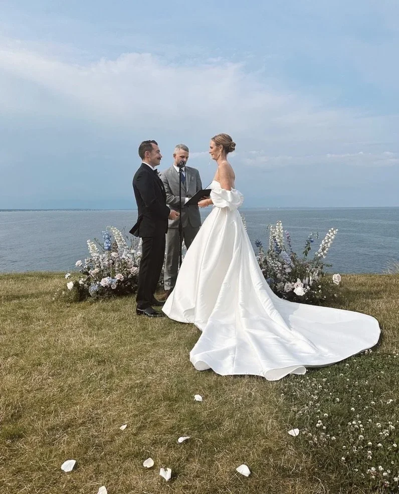 A wedding ceremony taking place outdoors near a body of water, with a bride in a white gown, a groom in a black suit, and an officiant, surrounded by flowers.