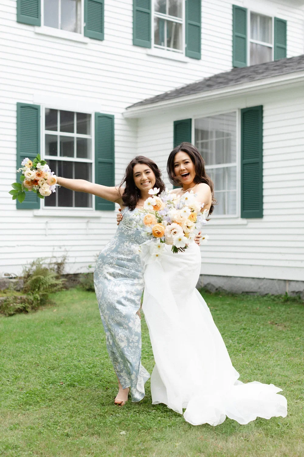 Two women in wedding dresses holding bouquets, smiling and celebrating outdoors in front of a white house with green shutters.