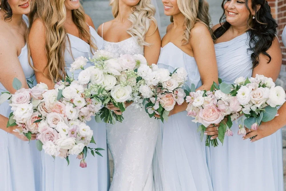 Four women in light blue dresses holding large bouquets of white and pink flowers at a wedding.