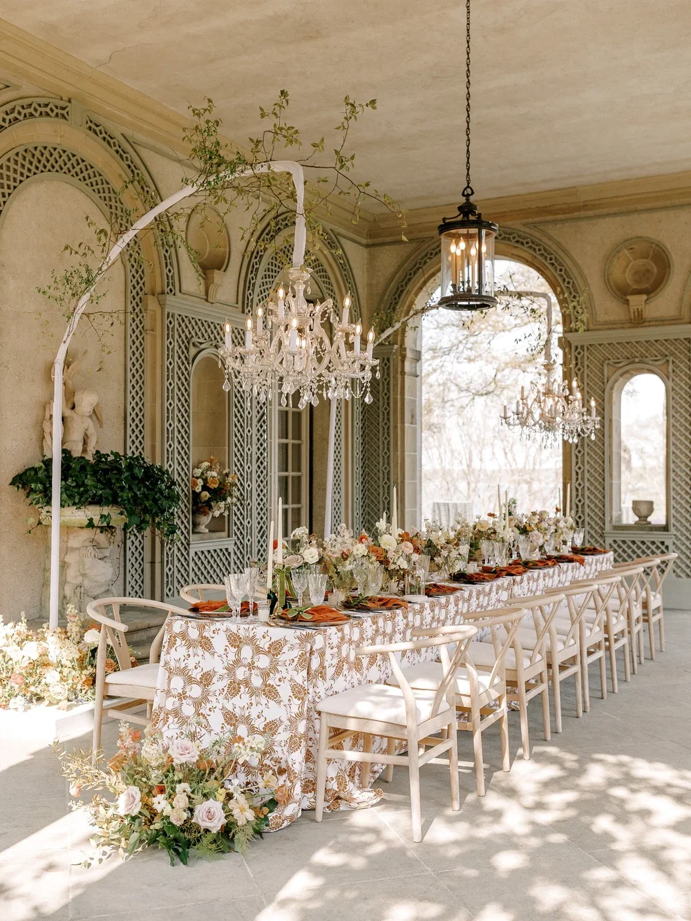 Elegant outdoor dining table set for a special occasion with floral arrangements, candles, and crystal glassware, under chandeliers and lantern lights, on a covered porch with ornate lattice walls.