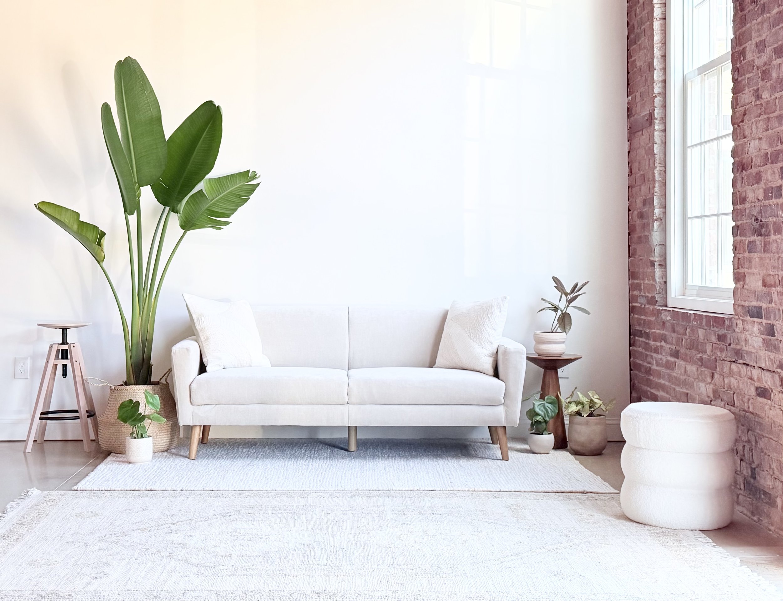 Minimalist living room with a white sofa, green indoor plants, a woven basket, a small side table, and a white ottoman, with a brick wall and large windows.