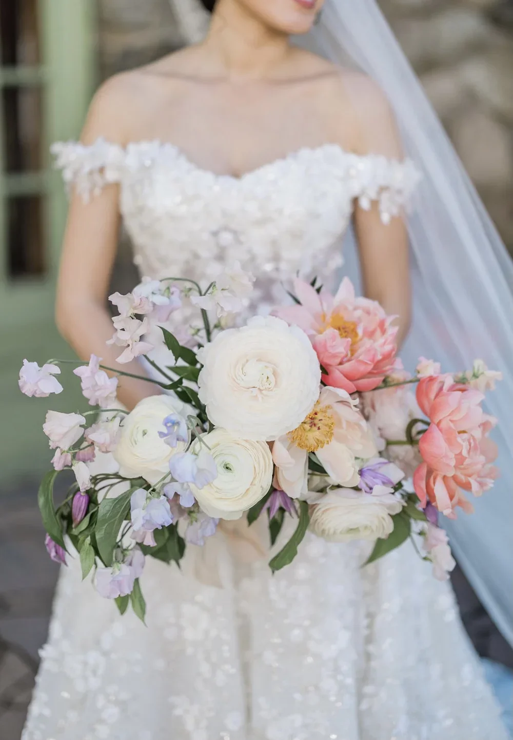Bride holding a bouquet of white, pink, and purple flowers in a wedding dress with off-shoulder lace details.