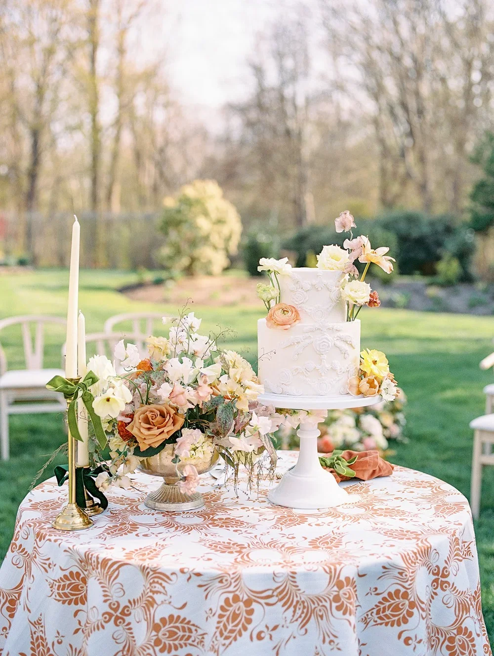 A white wedding cake decorated with pastel-colored flowers on a white cake stand, set on an outdoor table with a floral tablecloth and a floral centerpiece, surrounded by chairs and trees in the background.