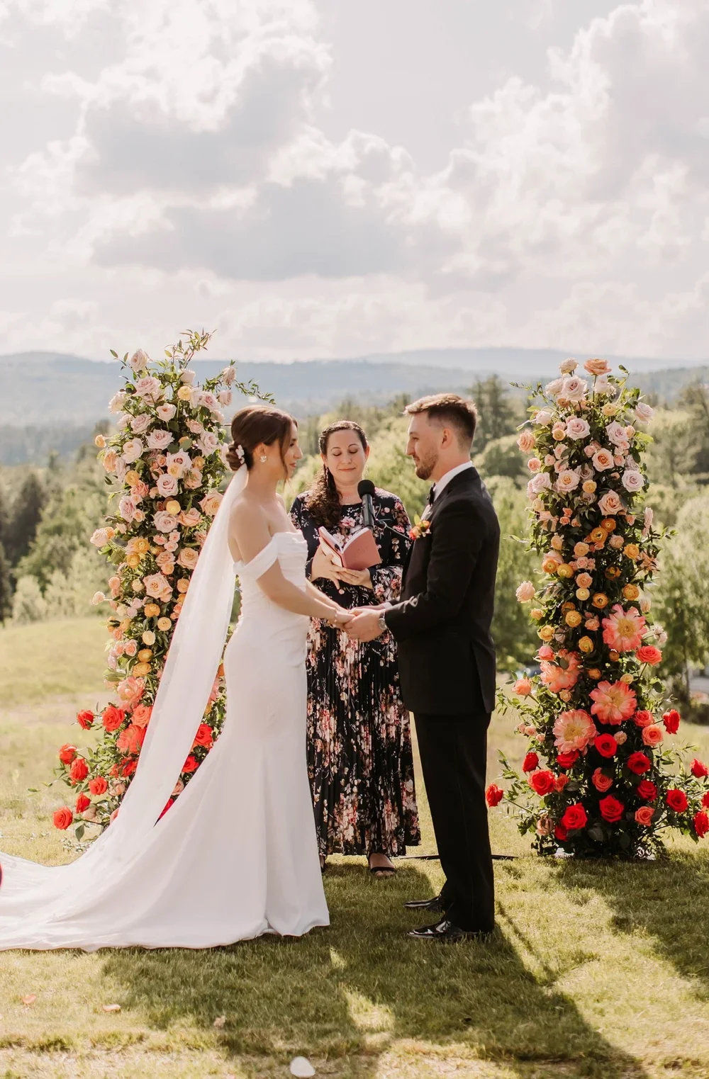 A bride and groom holding hands during an outdoor wedding ceremony, with a female officiant in the middle, surrounded by large floral arrangements of pink and red roses.