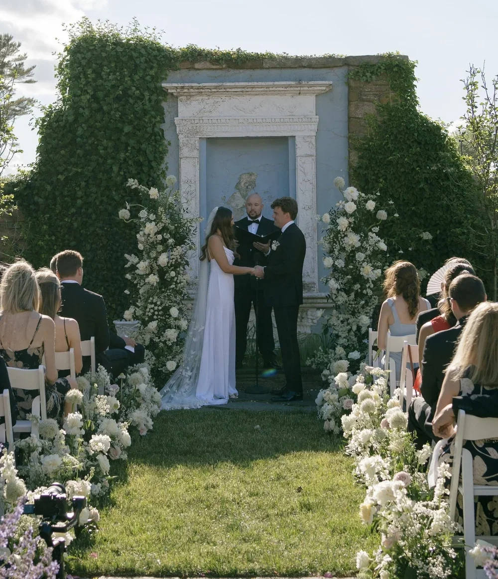 A wedding ceremony outdoors with a bride and groom exchanging vows in front of an officiant, surrounded by guests seated on white chairs decorated with white flowers, and a backdrop of greenery and an ancient stone wall.