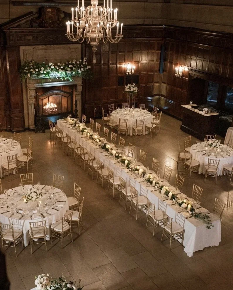 Elegant wedding reception with round tables and a long head table decorated with white flowers, in a wood-paneled hall with a chandelier and a fireplace adorned with flowers.