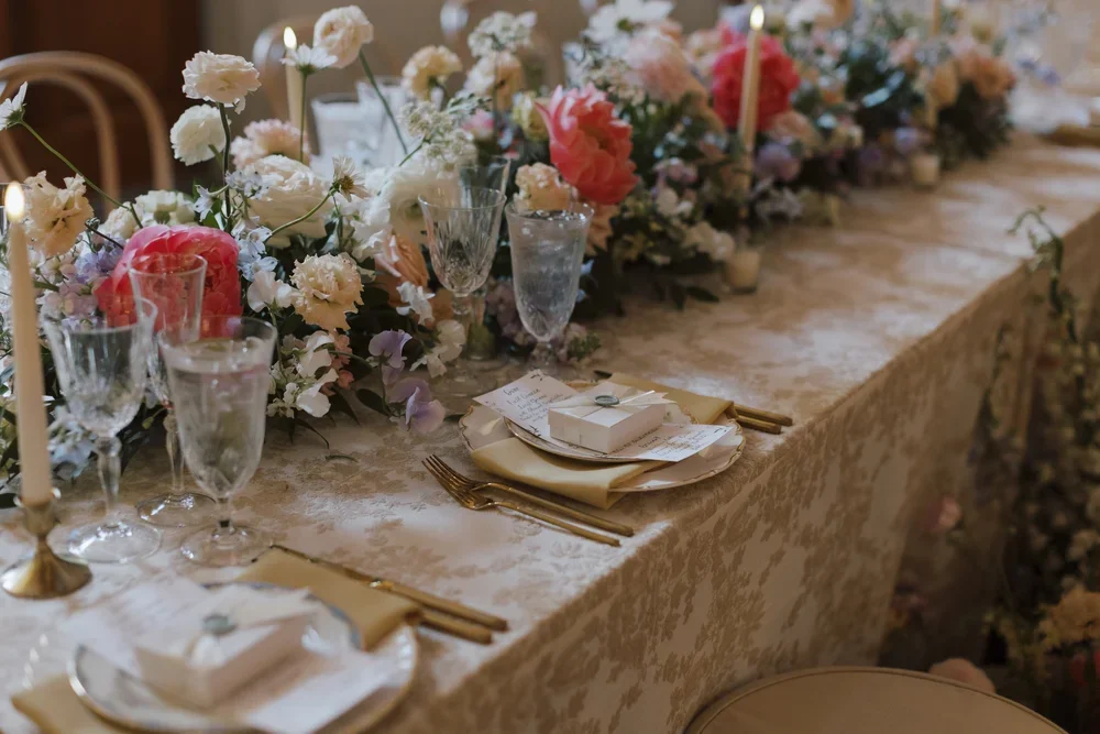 Elegant banquet table decorated with a floral centerpiece, candles, glassware, gold cutlery, and place settings with napkins and small boxes.