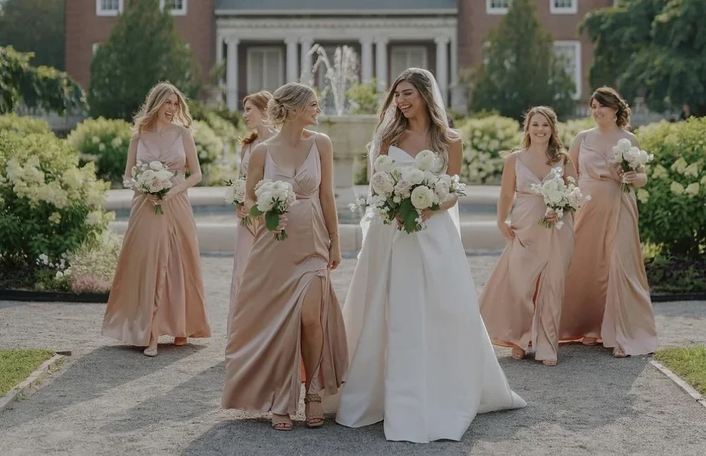 A bride and five bridesmaids walking outside near a fountain, holding bouquets of white flowers, with a large house in the background.