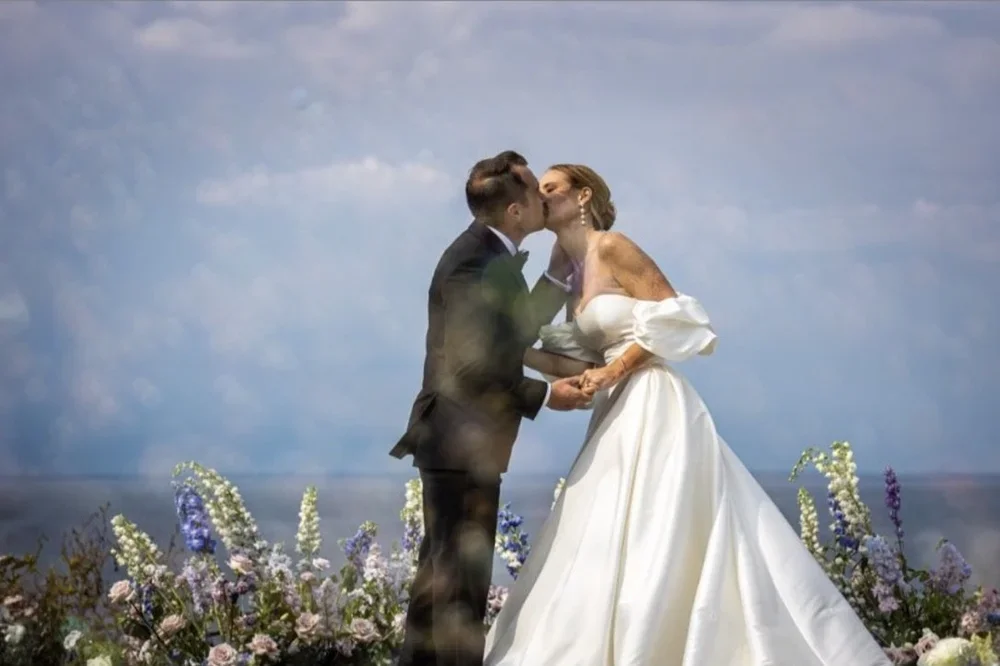 A bride and groom kissing outdoors on a wedding day, holding hands, surrounded by purple and white flowers, with a cloudy sky in the background.