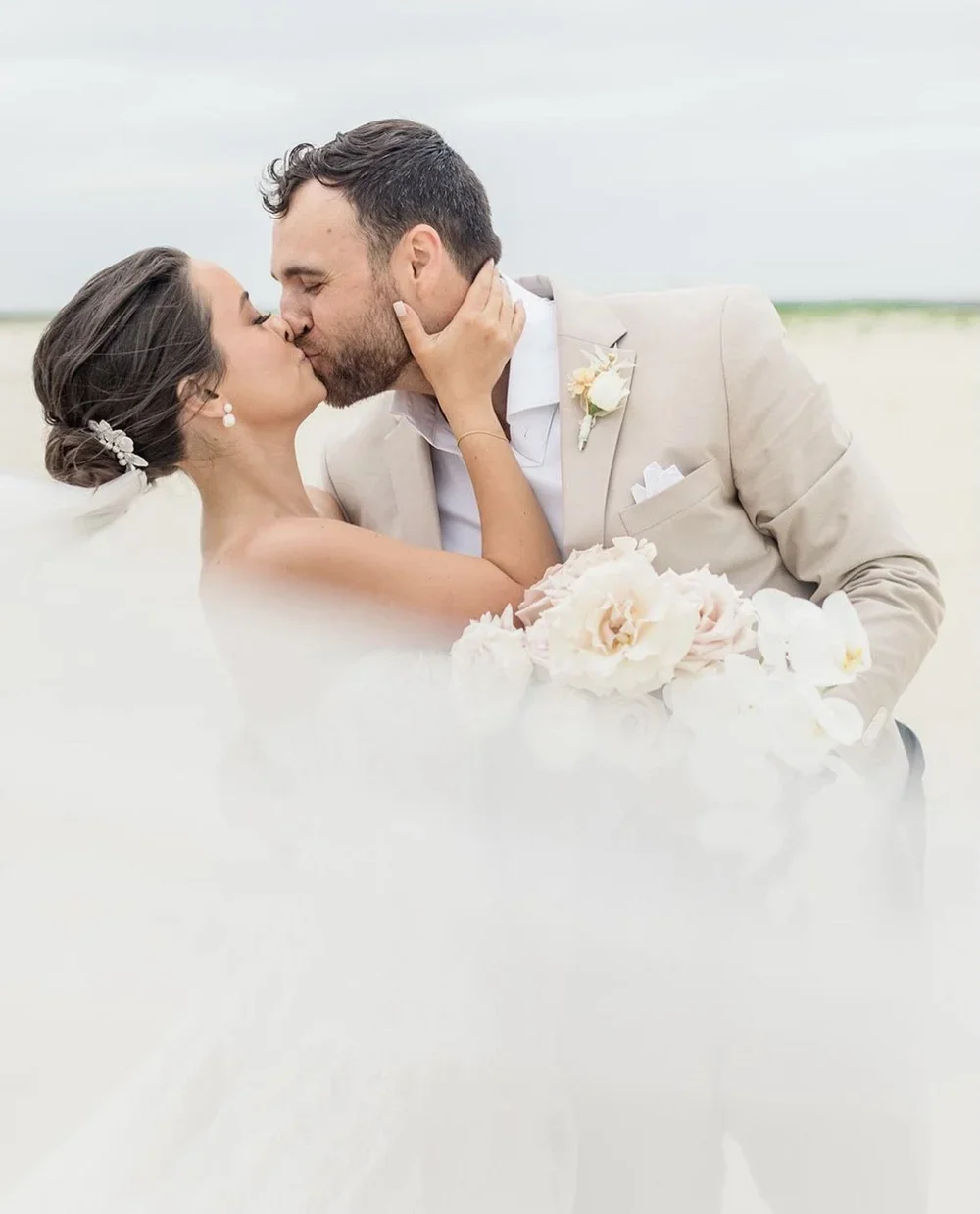 A bride and groom kiss on a beach during their wedding, with the bride holding a bouquet of white and blush flowers.