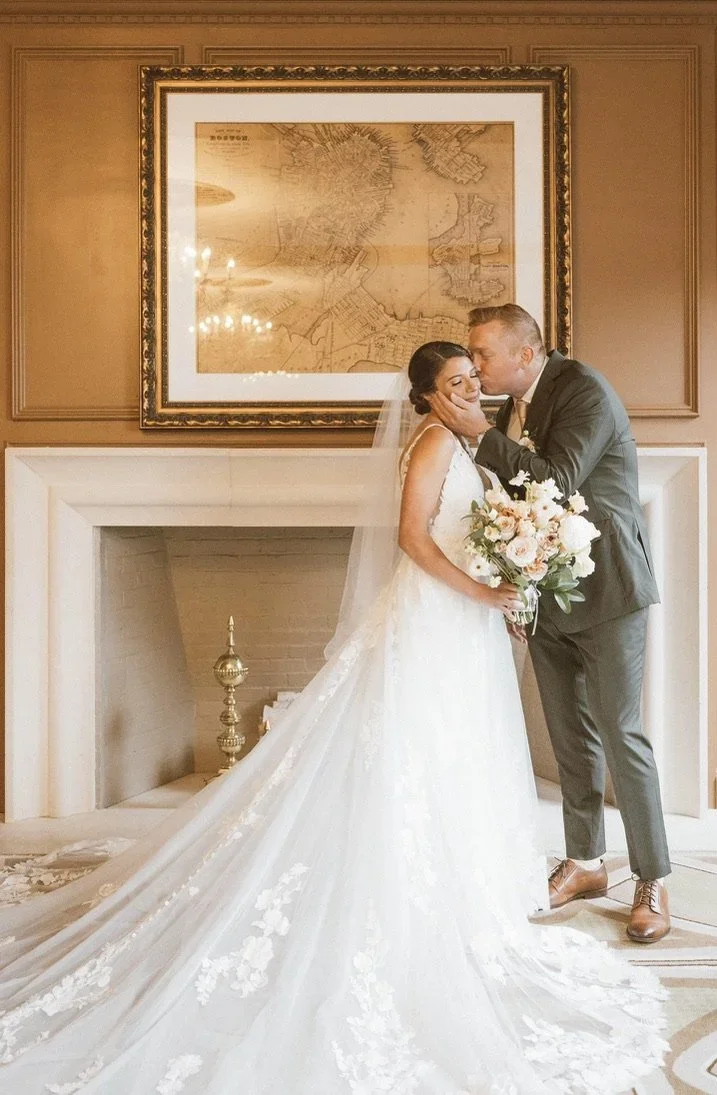 Wedding couple sharing a kiss indoors, woman in a white wedding dress holding a bouquet of flowers, man in a gray suit, large framed map on the wall behind them.
