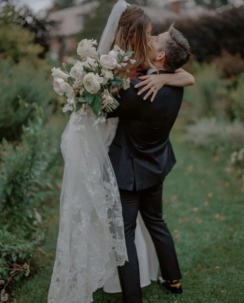 A bride in a white wedding gown holding a bouquet of white flowers is being lifted by a groom in a black tuxedo, both sharing a kiss outdoors in a garden.