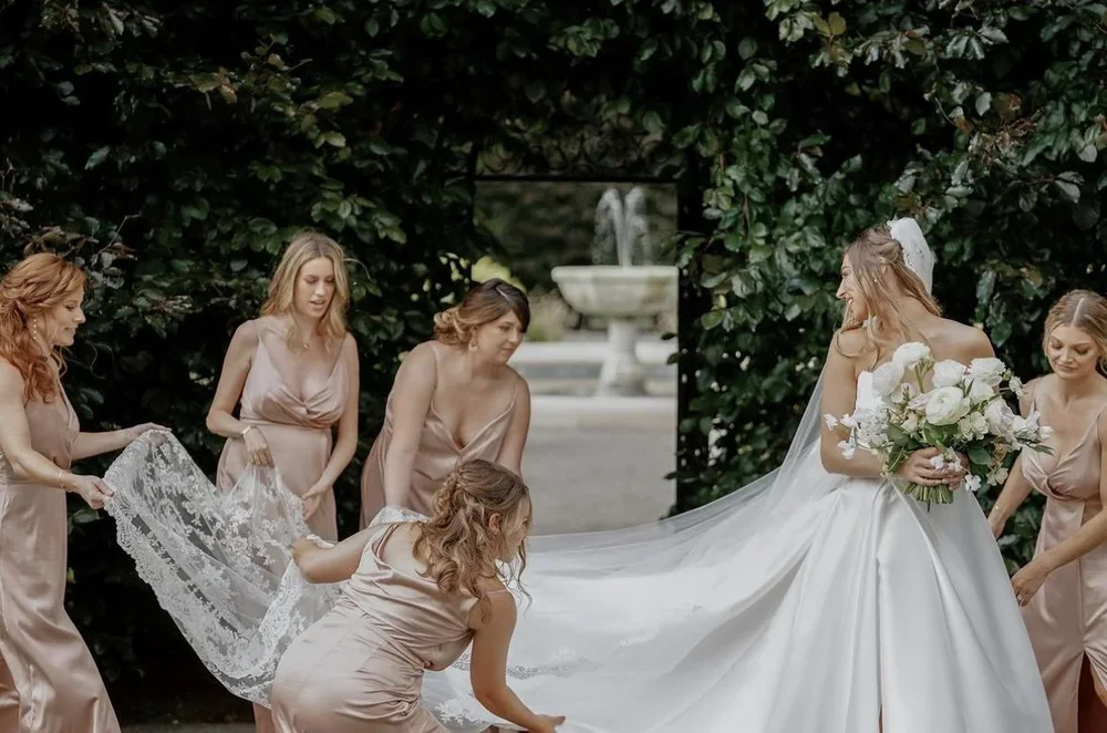 Bride with bouquet of white and pink flowers standing in a lush green garden, surrounded by her bridesmaids assisting with her wedding dress.