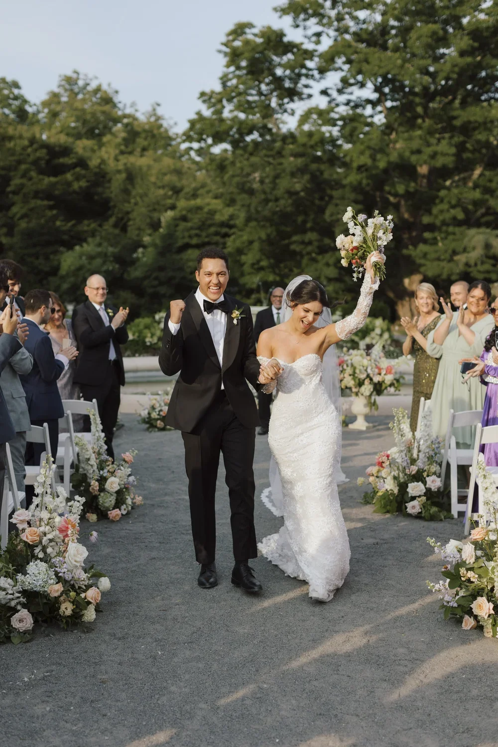 Bride and groom celebrating after wedding ceremony, walking down aisle with guests clapping and smiling, outdoor setting with flowers and trees.