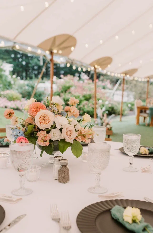 A table decorated with a pink and white floral centerpiece, surrounded by glassware and black plates, set up outdoors under a large white canopy with string lights and greenery in the background.