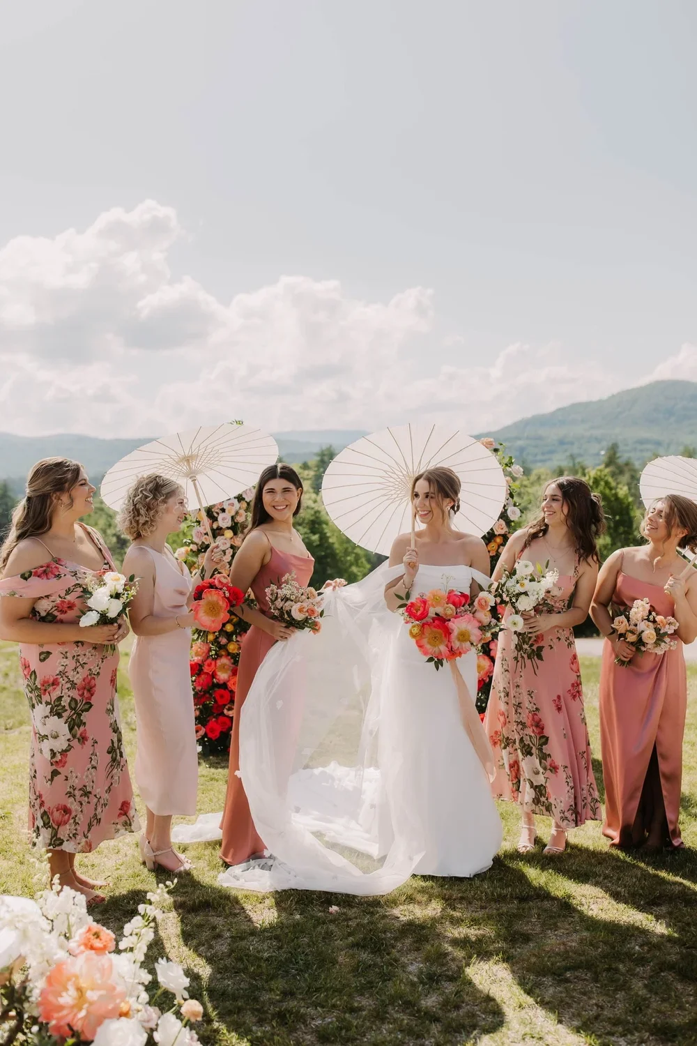 A group of six women, including a bride in a white wedding dress, holding bouquets of flowers and parasols, smiling outdoors during a wedding ceremony with a scenic mountain backdrop.