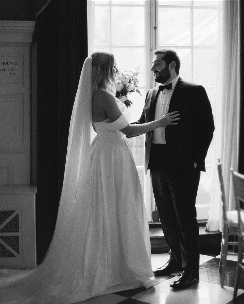 A bride and groom sharing a moment indoors by a window, with the bride in a wedding dress and veil, and the groom in a tuxedo, smiling at each other.