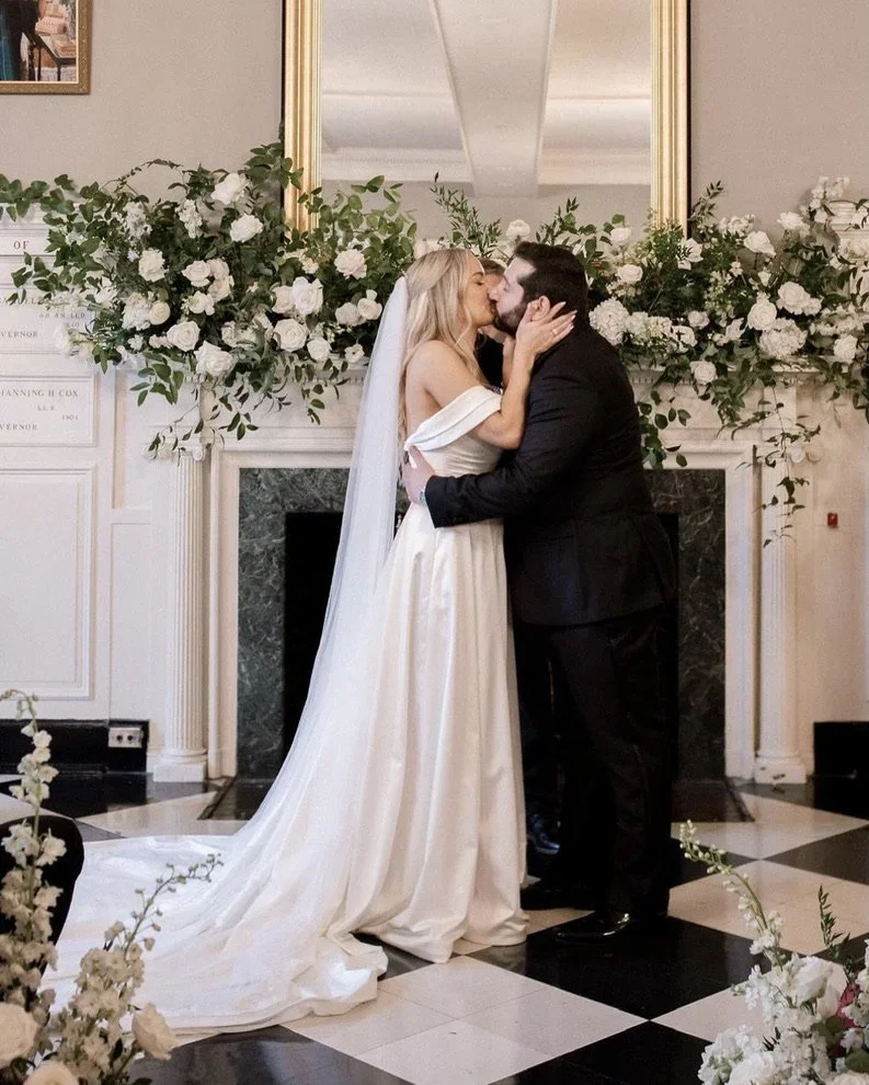 A bride and groom share a kiss at their wedding in front of a flower arrangement and a mirror.
