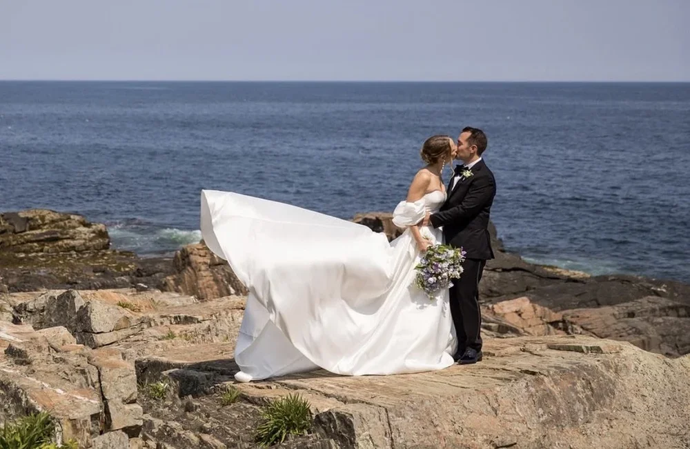 A bride and groom kissing on a rocky coastline, with the ocean in the background. The bride is holding a bouquet of flowers and wearing a white wedding dress, while the groom is dressed in a black tuxedo.