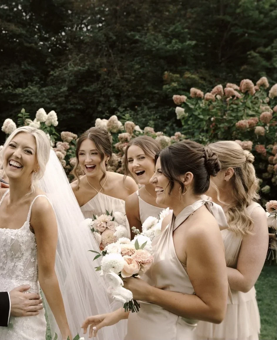 A bride and five bridesmaids smiling and laughing in an outdoor garden setting with pink and white flowers.