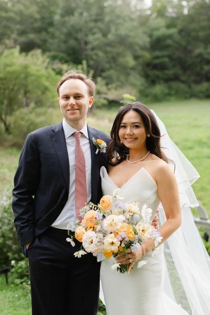 A bride and groom standing outdoors on a green lawn with trees in the background, smiling for a wedding photo. The bride is holding a bouquet of pastel flowers, wearing a strapless white wedding dress and a pearl necklace. The groom is dressed in a d
