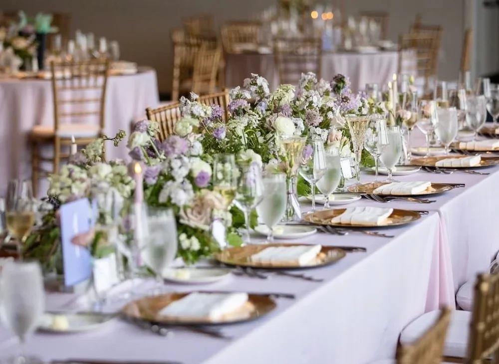 Elegant banquet table decorated with pastel floral arrangements, glassware, gold chargers, and white napkins in a decorated event space