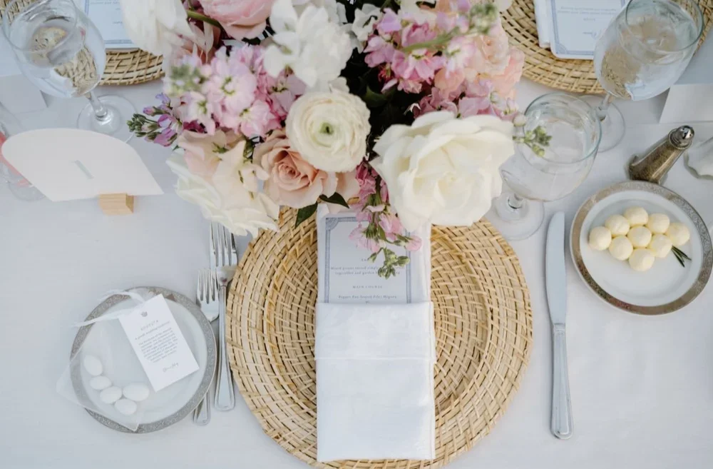 Elegant table setting with a floral centerpiece, woven placemat, white napkin, silverware, glassware, and small plates with white chocolates.