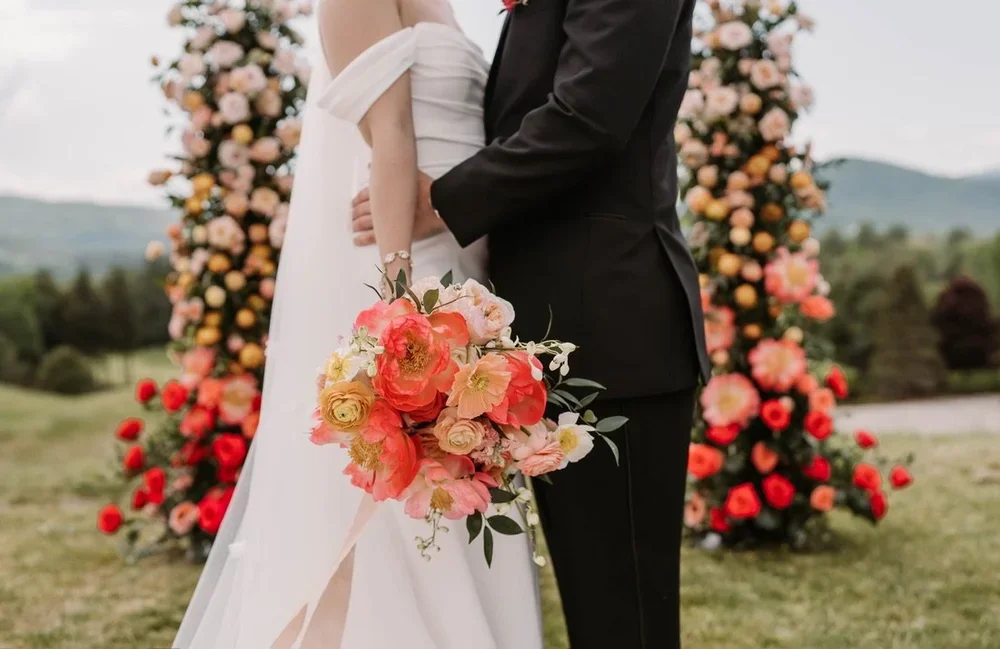 A bride and groom standing outdoors in front of a floral arch at a wedding, holding a colorful bouquet of flowers.