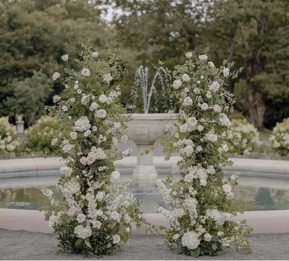 Floral arch made of white and pale pink roses and greenery, placed in front of a decorative fountain in a garden setting.