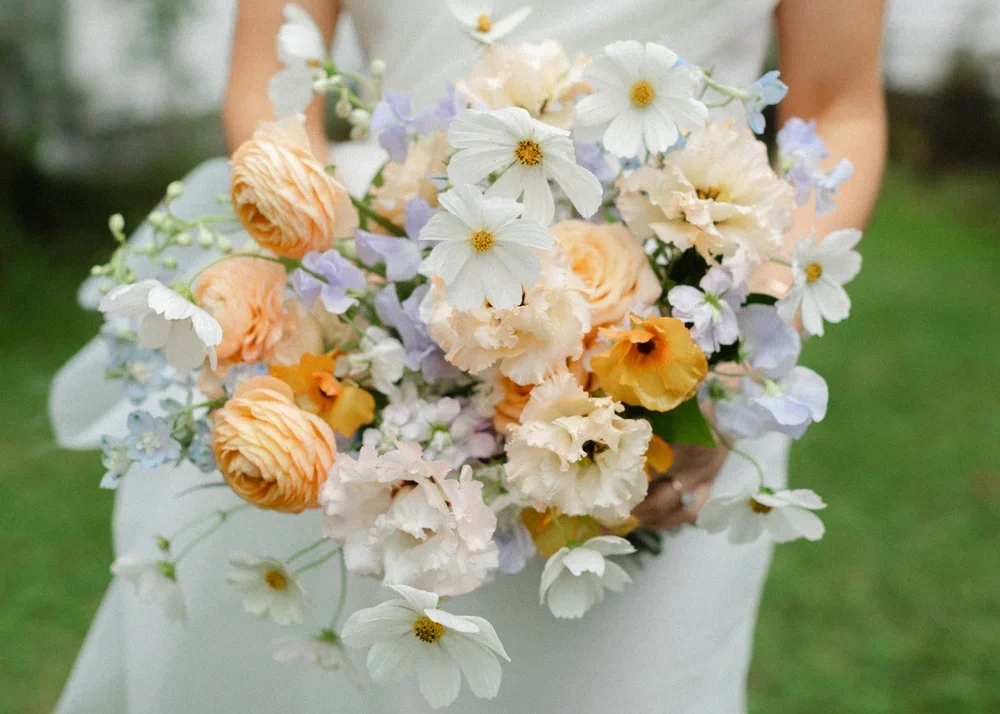 A person in a wedding dress holding a bouquet of peach, white, and purple flowers outdoors.