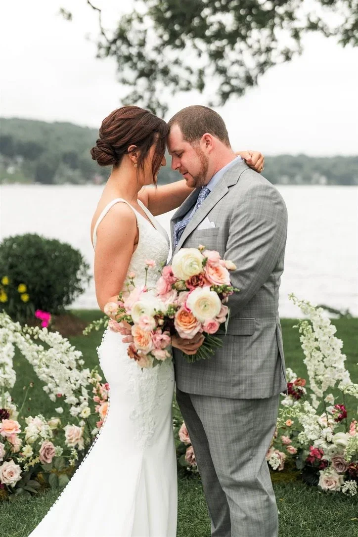 A bride and groom embrace during their wedding outdoors by a lake, with trees and floral arrangements in the background.