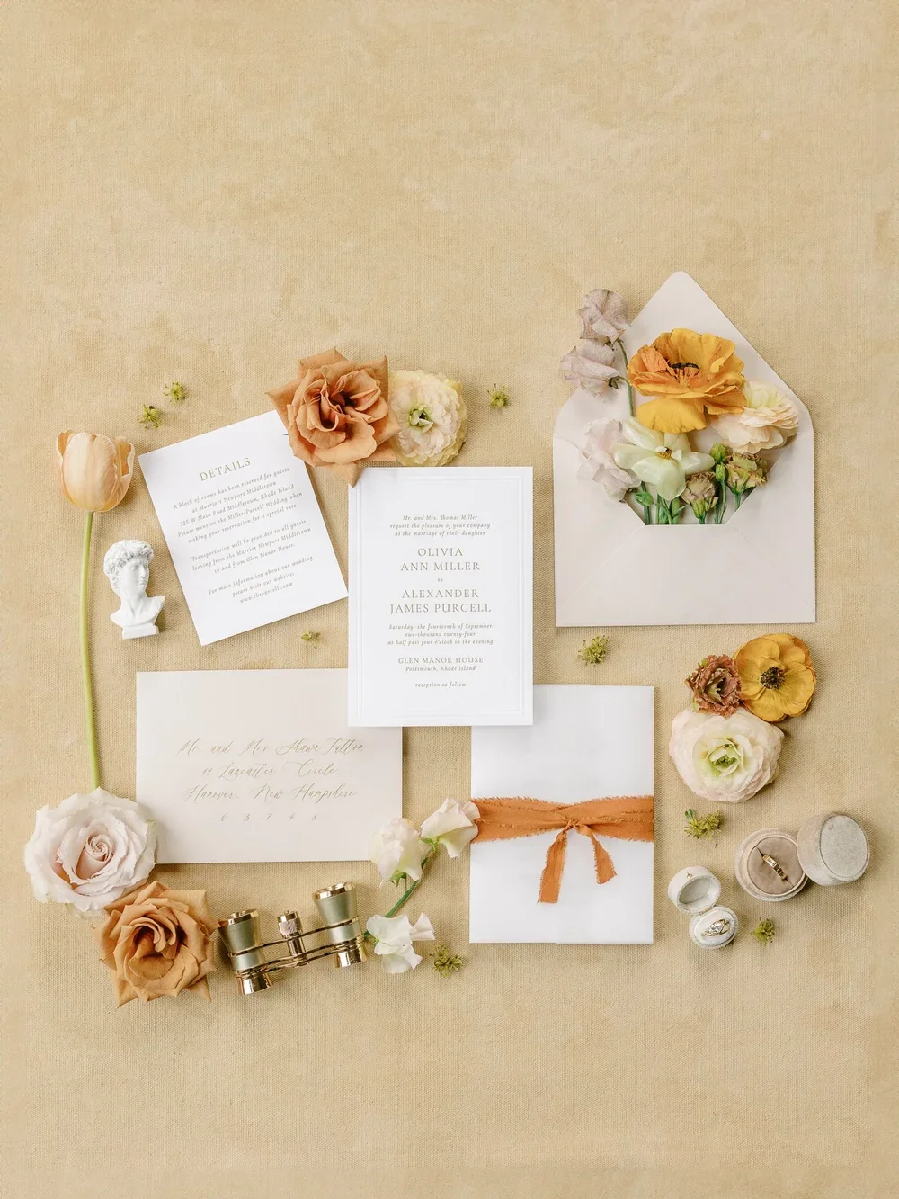 Flat lay of wedding invitation, details card, envelopes with flowers, ring box, binoculars, and small decorative bust, all arranged on a beige background.