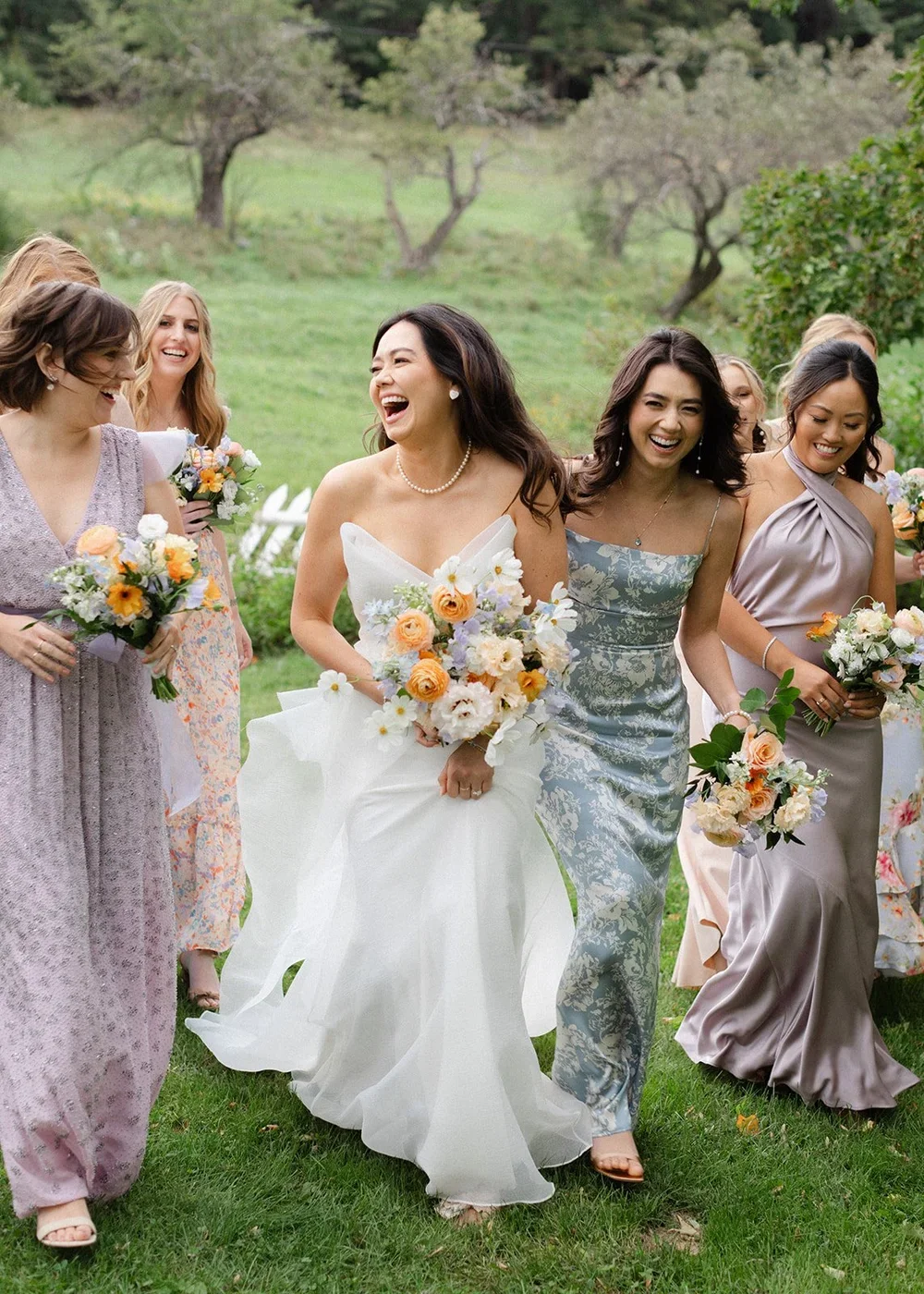A group of women, including a bride in a white wedding dress, walking outdoors with joyful expressions, holding bouquets of flowers, on a grassy area with trees in the background.