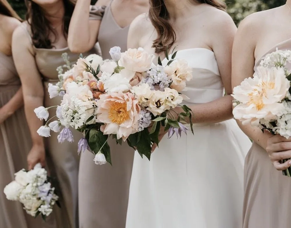 Bride holding a bouquet of pastel-colored flowers during a wedding, surrounded by bridesmaids in beige dresses, outdoors.