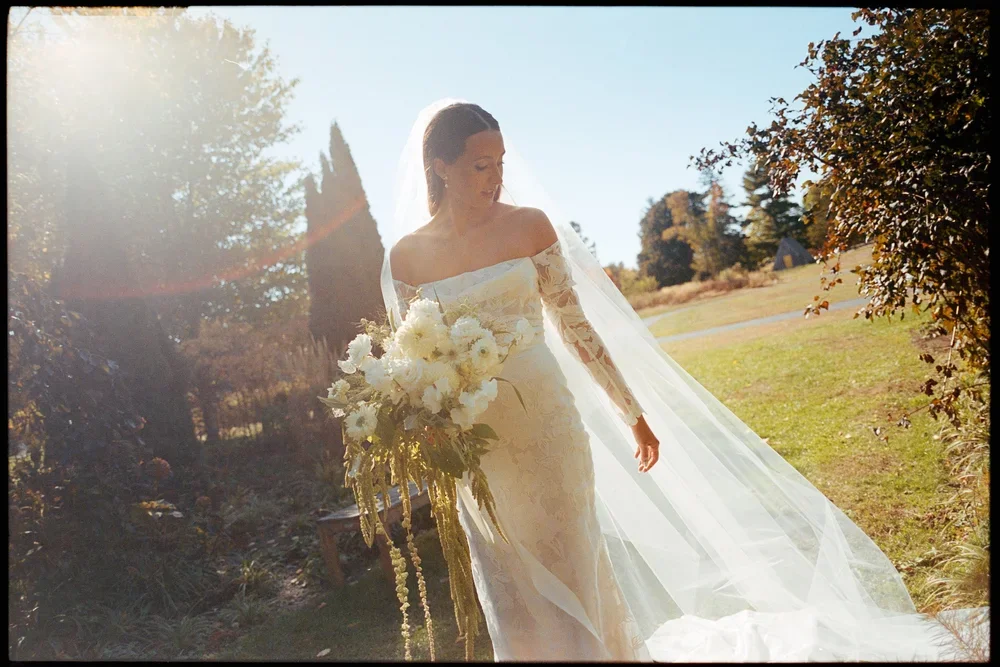 A bride in a long-sleeved lace wedding gown with an off-shoulder neckline holding a large bouquet of white flowers outdoors during sunset.