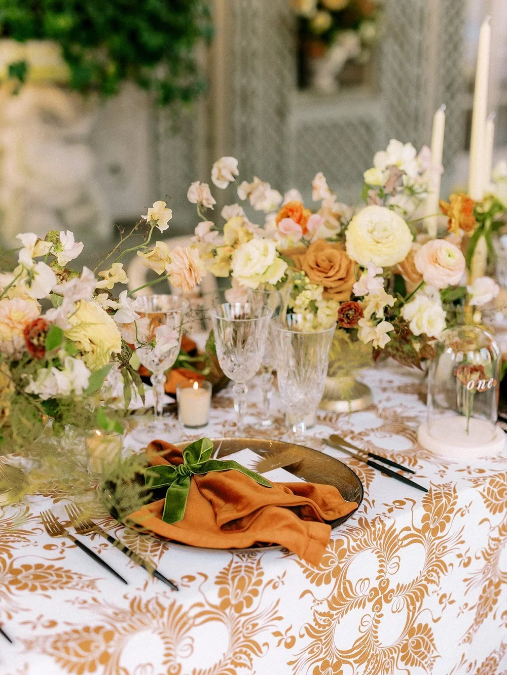 Elegant dining table with floral centerpiece, glassware, candles, and table settings, featuring orange and peach napkins with green ribbon, set outdoors.