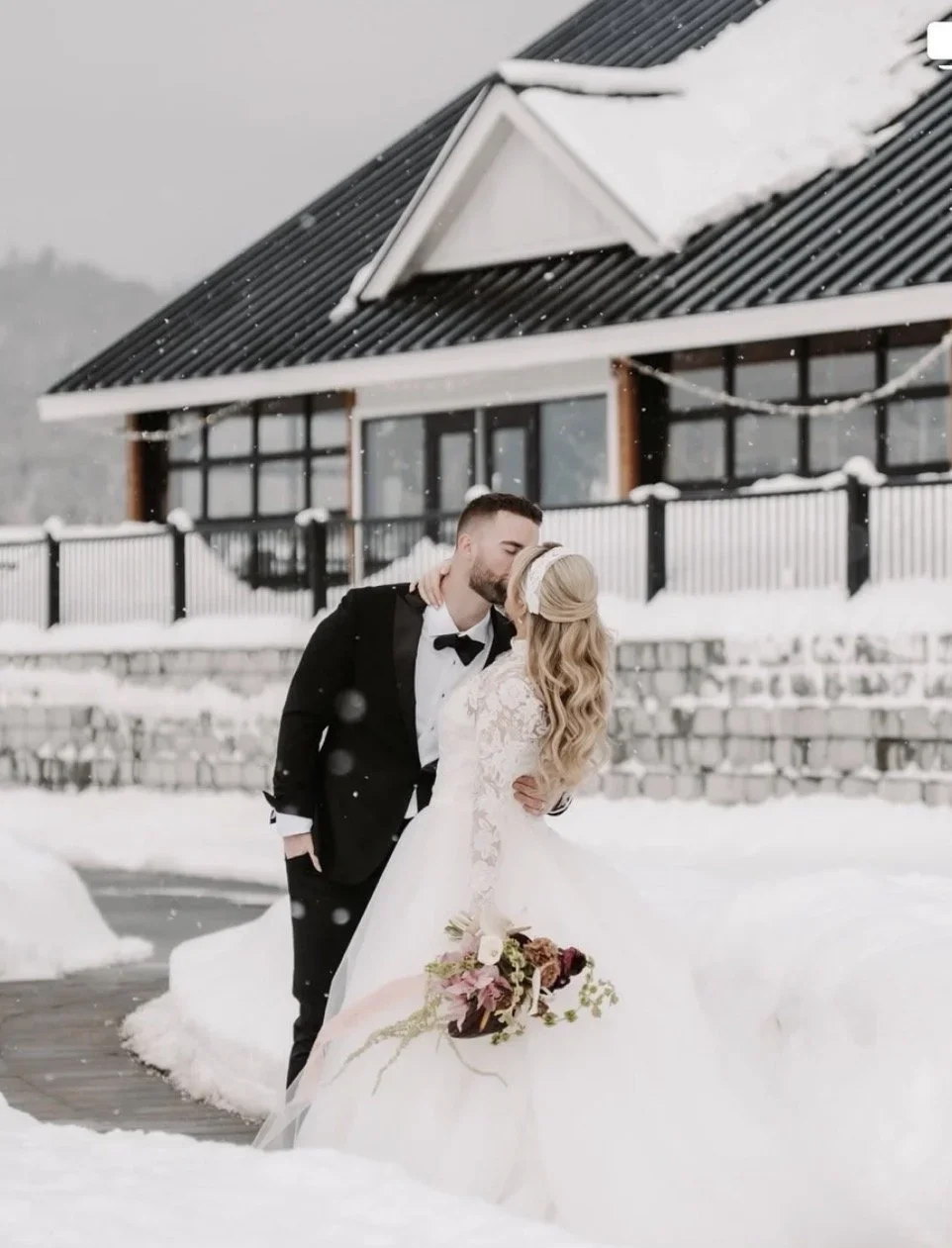 A bride and groom in wedding attire sharing a kiss outdoors in a snowy landscape, with a house and a stone wall in the background.