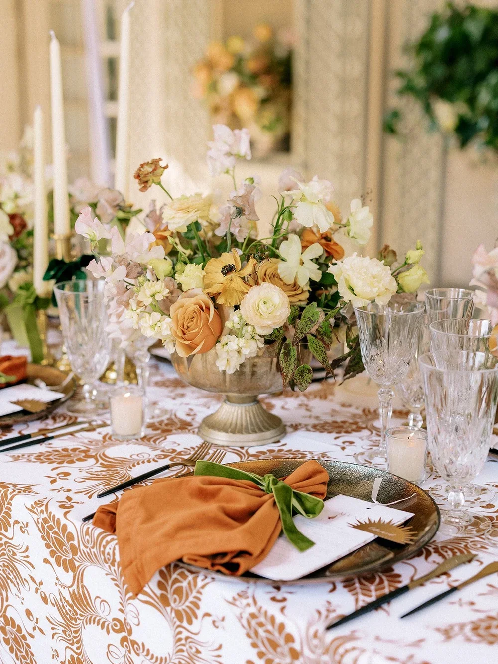 Elegant table setting with a floral centerpiece, candlesticks, glassware, and a cloth napkin tied with a green ribbon on a patterned tablecloth.