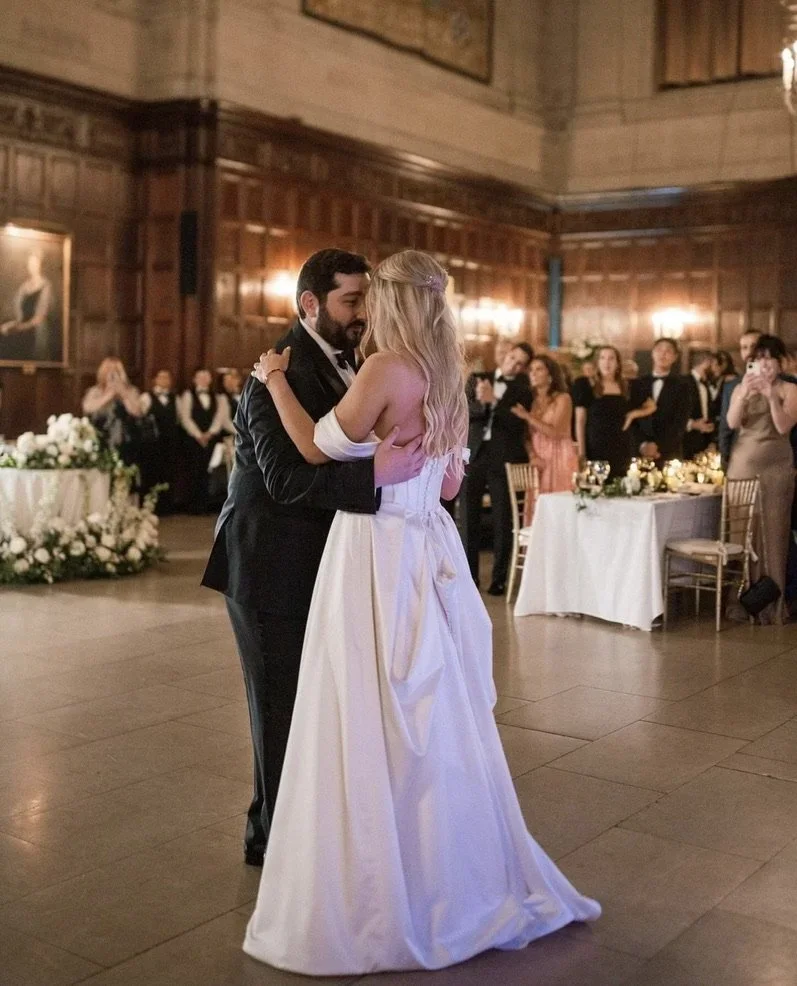 A bride and groom are sharing their first dance at a wedding reception in a decorated banquet hall with guests watching.