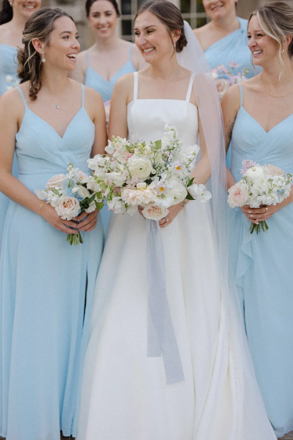 Bride in a white wedding gown holding a bouquet of flowers surrounded by bridesmaids in light blue dresses holding smaller bouquets at an outdoor wedding ceremony.