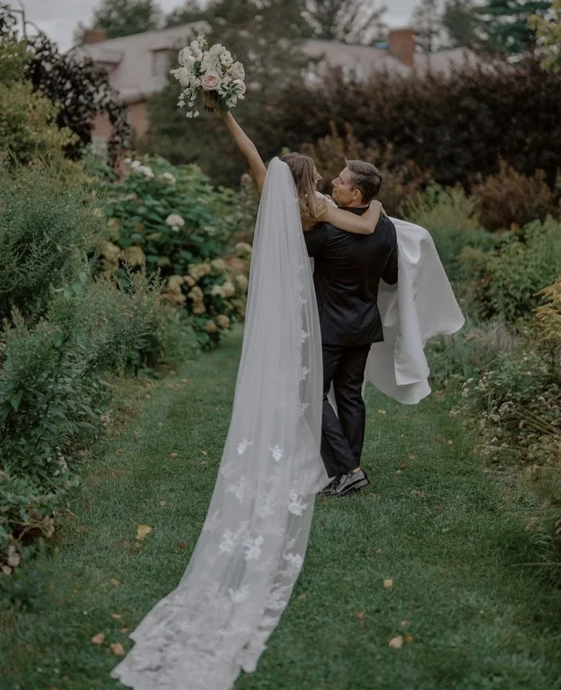 A bride in a wedding dress with a long veil is being lifted by the groom, holding a bouquet, in a garden setting.